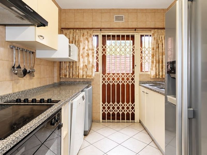 Functional kitchen featuring granite countertops, built-in oven, electric hob, extractor fan, and tiled finishes with direct access to the exterior via a security gate.