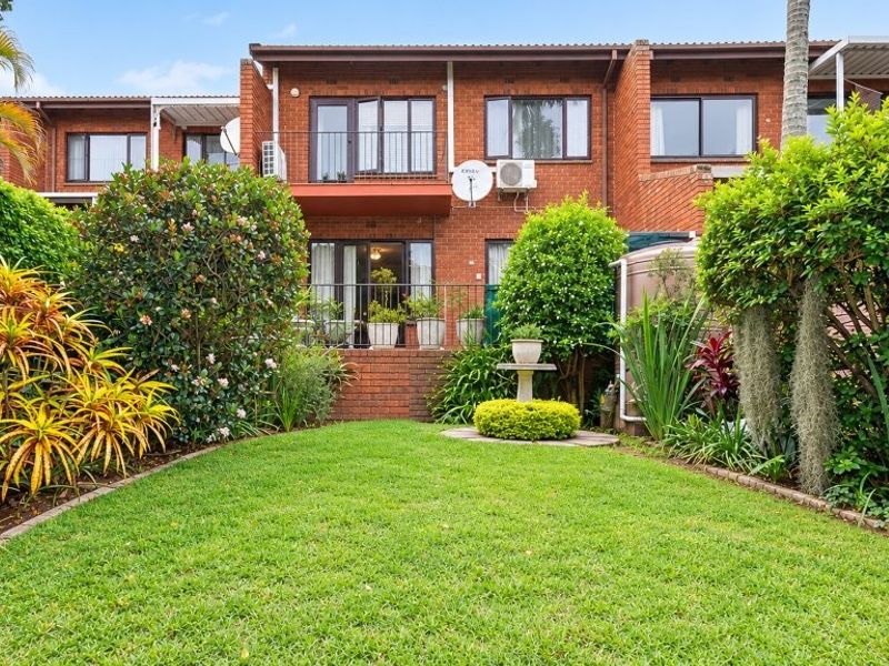 Rear view of a brick townhouse featuring a private garden, balcony, and established landscaping.