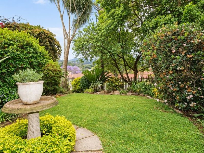 A well-maintained private garden featuring a lush green lawn, mature trees, established shrubbery, and a secure metal perimeter fence.