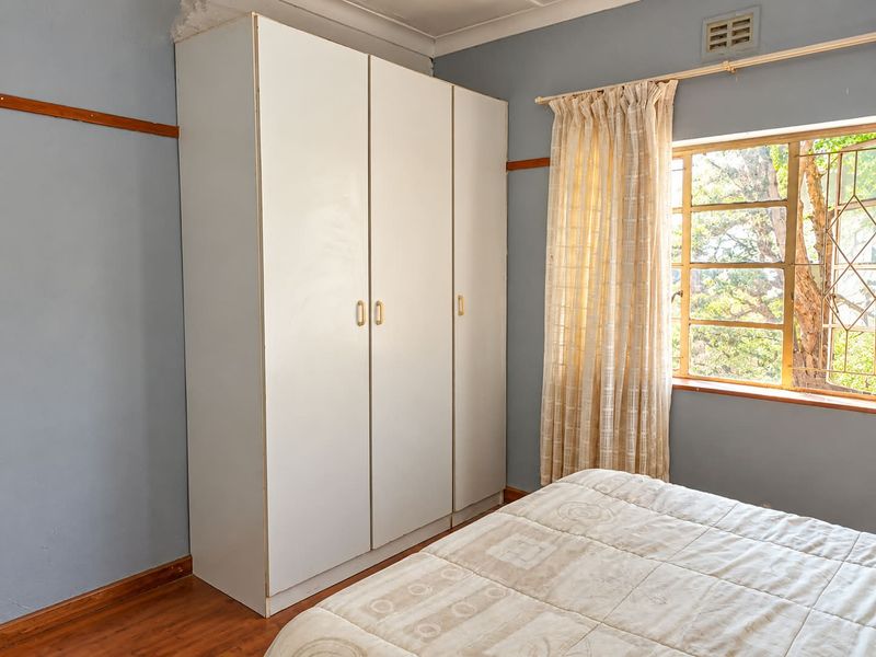 A bedroom featuring a built-in wardrobe, laminate flooring, and a steel-framed window with security bars, showing signs of wear on the ceiling and walls.