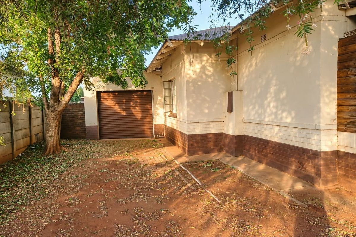 Rear exterior view of the property featuring a roller garage door, brick boundary wall, and plastered masonry walls with mature tree coverage.