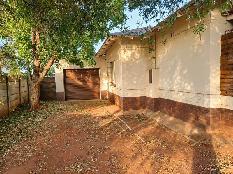 Rear exterior view of the property featuring a roller garage door, brick boundary wall, and plastered masonry walls with mature tree coverage.