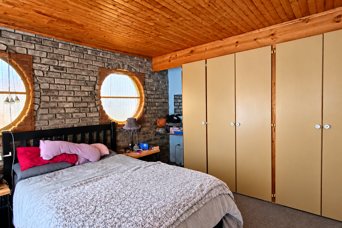 Bedroom featuring a distinctive exposed brick wall, circular windows, and a warm timber ceiling with ample built-in wardrobe storage.