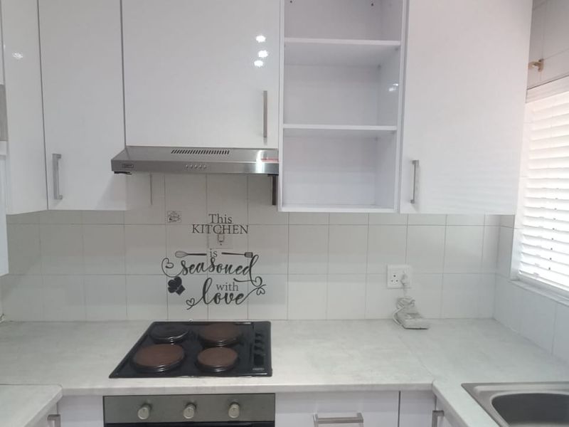 A clean, functional kitchen featuring white cabinetry, a built-in electric hob, oven, extractor fan, and a stainless steel sink with tiled backsplash.