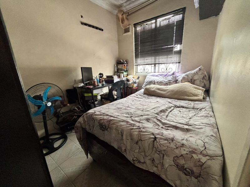 A compact bedroom featuring tiled flooring, a window with security bars and venetian blinds, and visible wall and ceiling wear requiring maintenance.