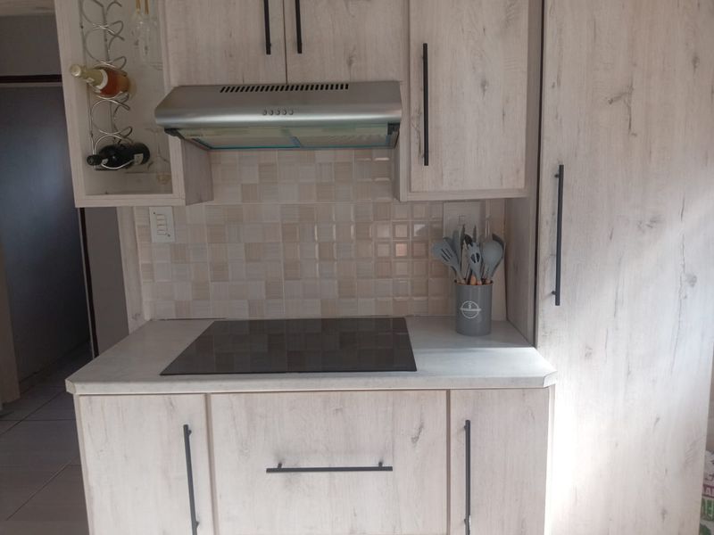 A modern kitchen area featuring light wood-grain cabinetry, a sleek electric hob, a stainless steel extractor fan, and a neutral tiled backsplash.