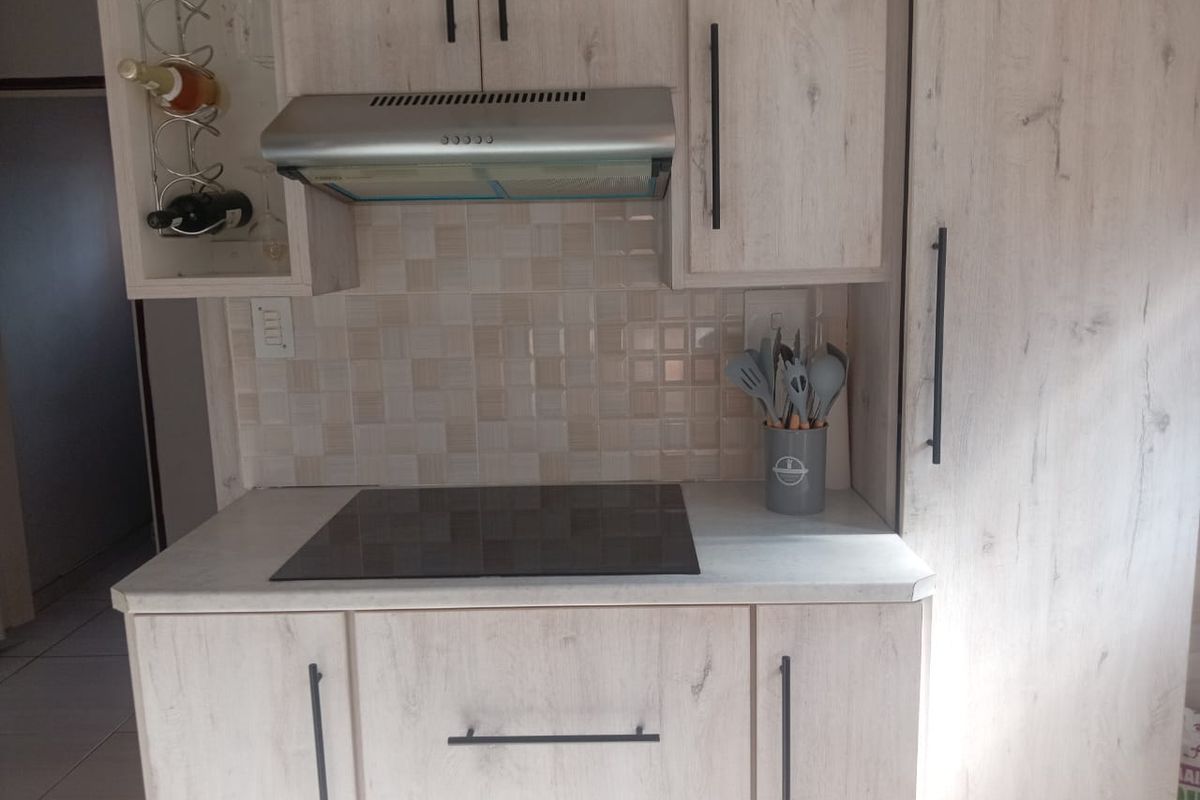 A modern kitchen area featuring light wood-grain cabinetry, a sleek electric hob, a stainless steel extractor fan, and a neutral tiled backsplash.