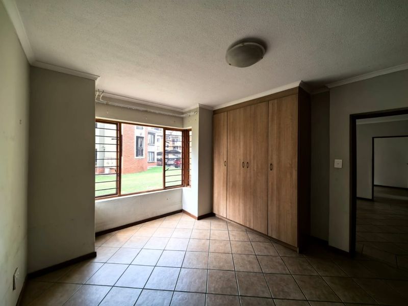 A vacant bedroom featuring tiled flooring, built-in wooden cupboards, and large windows with security bars, offering a functional and neutral living space.