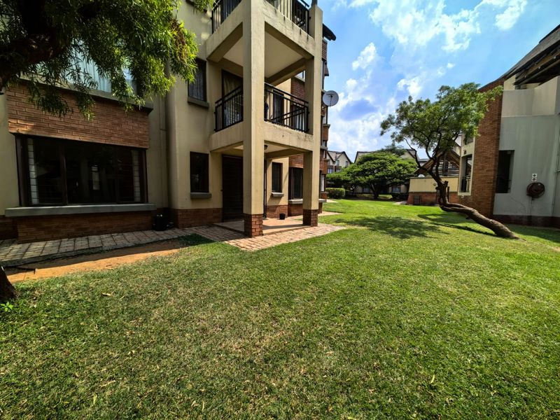 Rear view of the apartment building featuring a brick facade, private balcony, paved patio area, and well-maintained communal garden space.