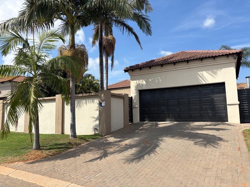 Front exterior view of the property featuring a paved driveway, double garage, and perimeter wall with established palm trees.