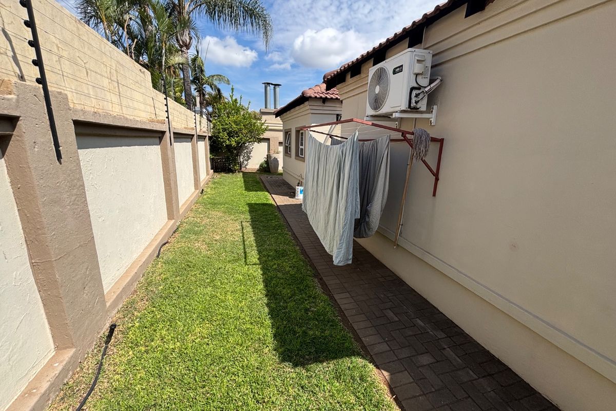 A well-maintained rear exterior area featuring a paved walkway, manicured lawn, perimeter wall with electric fencing, and a wall-mounted air conditioning unit.