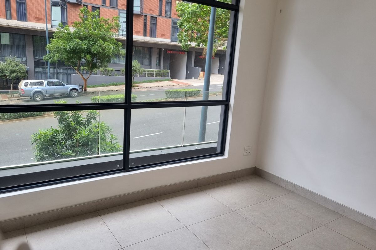 A bright, empty living area featuring large black-framed windows with street views and neutral tiled flooring.