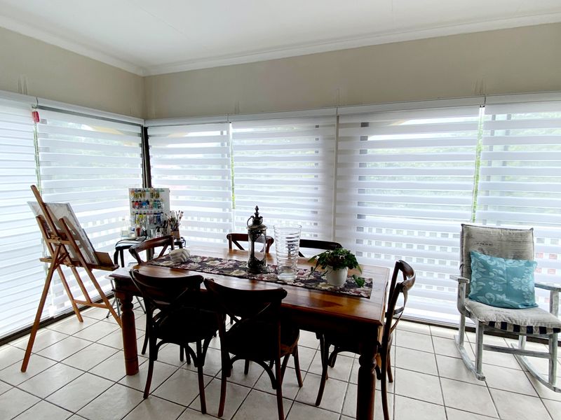 A bright dining area featuring large windows with modern dual-layer roller blinds and neutral tiled flooring.