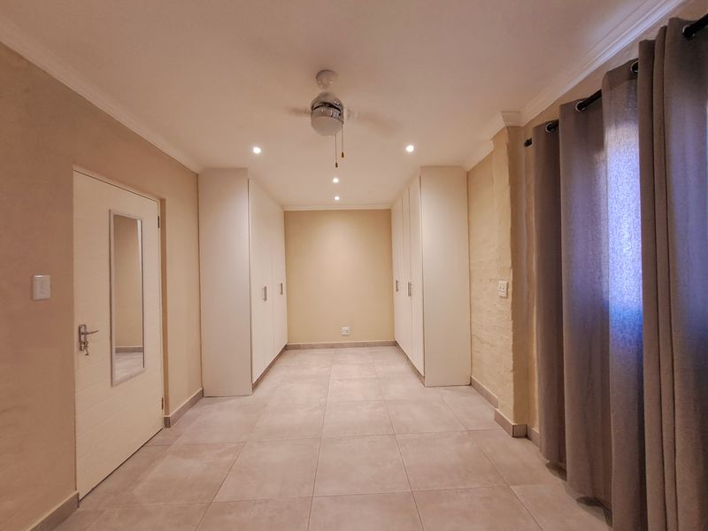 A spacious, empty bedroom featuring neutral tiled flooring, ample built-in cupboard storage, a ceiling fan, and recessed lighting.