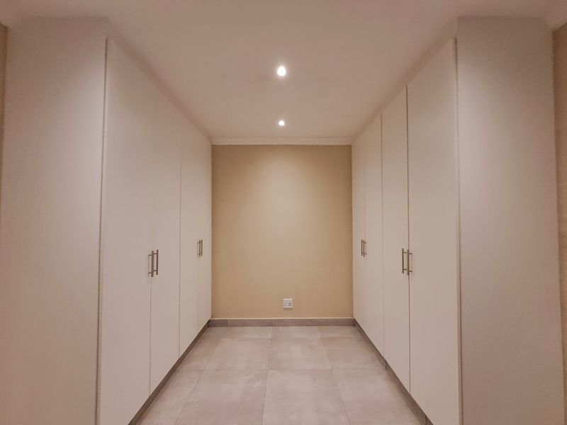 A clean and modern dressing area or bedroom space featuring extensive built-in cupboards, neutral tiled flooring, and recessed ceiling lighting.