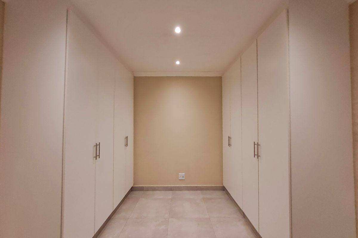 A clean and modern dressing area or bedroom space featuring extensive built-in cupboards, neutral tiled flooring, and recessed ceiling lighting.
