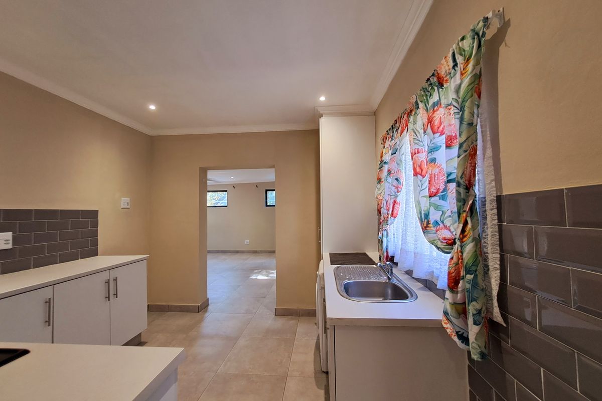A clean, functional kitchen featuring white cabinetry, stainless steel sink, grey tiled splashbacks, and neutral floor tiles, leading into an adjacent living area.