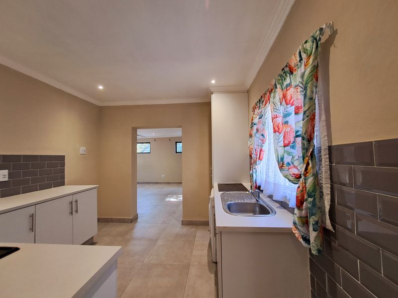 A clean, functional kitchen featuring white cabinetry, stainless steel sink, grey tiled splashbacks, and neutral floor tiles, leading into an adjacent living area.