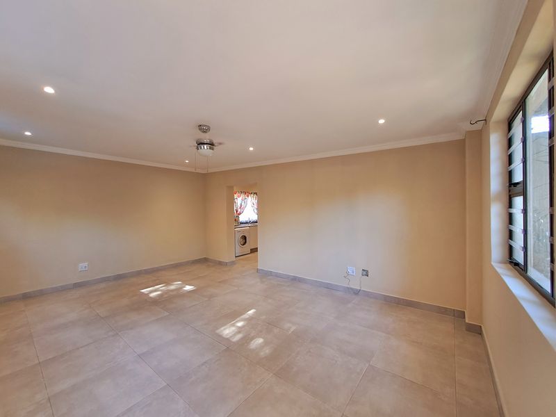 A spacious, empty living area featuring neutral tiled flooring, recessed lighting, a ceiling fan, and security bars on the windows.