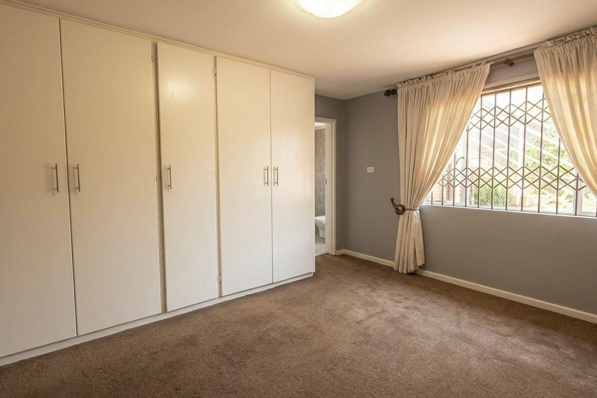 A spacious bedroom featuring neutral wall tones, carpeted flooring, and extensive built-in cupboard storage, complemented by a window with security bars.