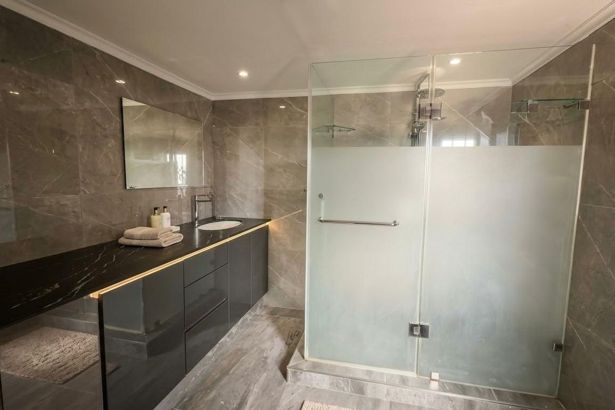 Modern bathroom featuring a sleek vanity with stone countertop, large wall mirror, and a spacious walk-in shower with glass enclosure and grey tiled finishes.
