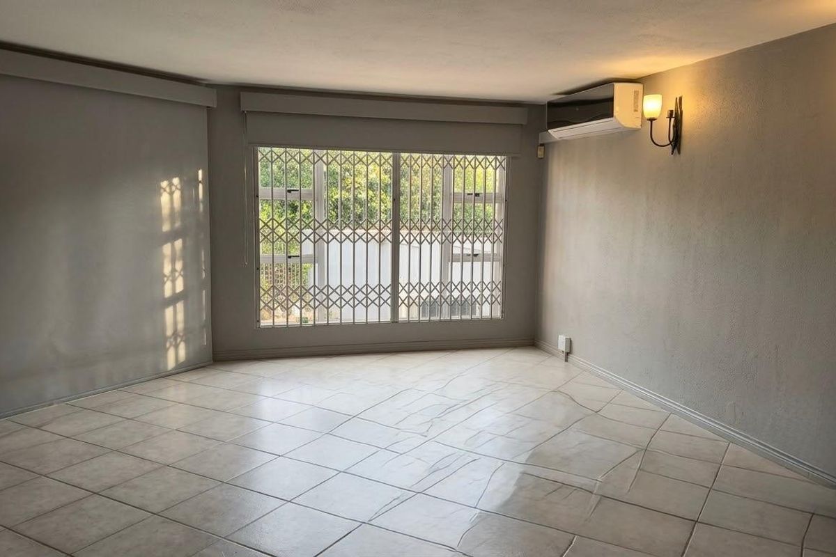A spacious, empty living area featuring neutral tiled flooring, wall-mounted air conditioning, and secure window treatments, offering a clean canvas for personalization.