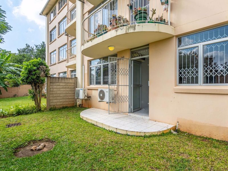 Rear view of the flat featuring a small tiled patio, security gates, and an upper-level balcony overlooking a shared garden area.