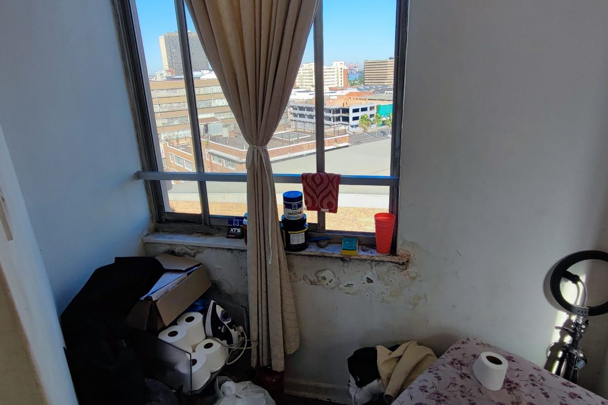 A bedroom featuring a large metal-framed window with urban views, showing significant wall damage and peeling paint beneath the sill.