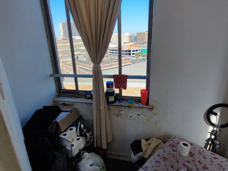 A bedroom featuring a large metal-framed window with urban views, showing significant wall damage and peeling paint beneath the sill.
