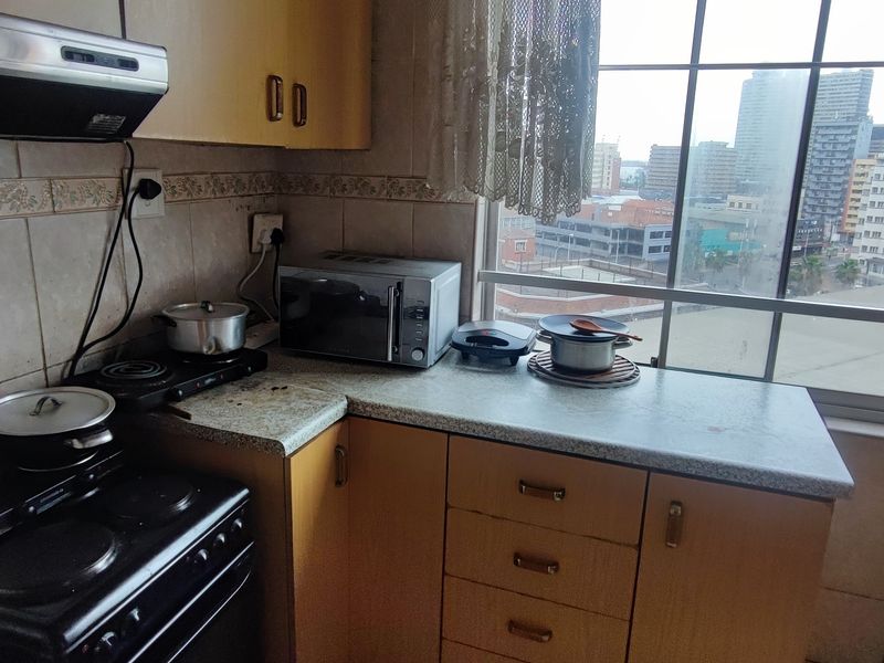 A compact kitchen featuring dated laminate cabinetry, tiled backsplash, and an extractor fan, with large windows offering an urban city view.