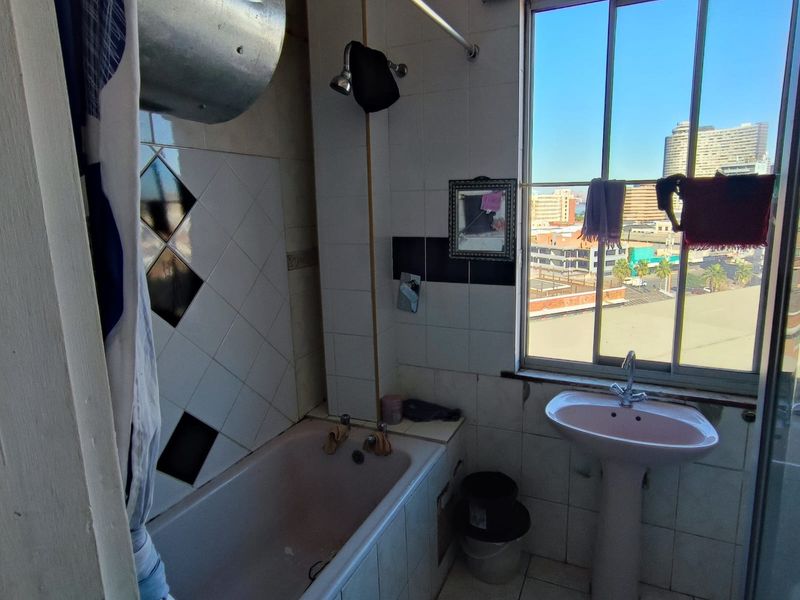A dated bathroom featuring a bathtub, pedestal basin, and tiled finishes, with a large window offering urban views.