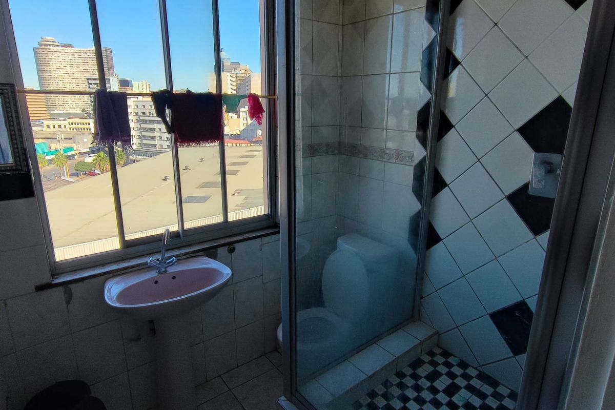 A dated bathroom featuring a pedestal basin, a glass-enclosed shower with patterned tiling, and a large window offering an urban city view.