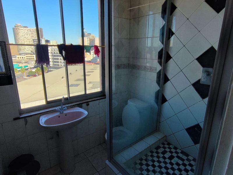 A dated bathroom featuring a pedestal basin, a glass-enclosed shower with patterned tiling, and a large window offering an urban city view.