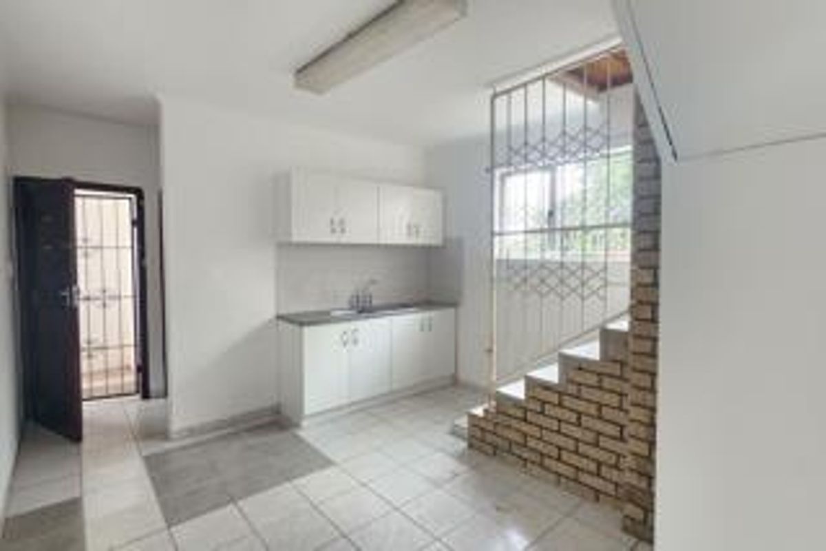 A compact, empty kitchen area featuring white built-in cabinetry, a stainless steel sink, tiled flooring, and a distinctive brick staircase feature.