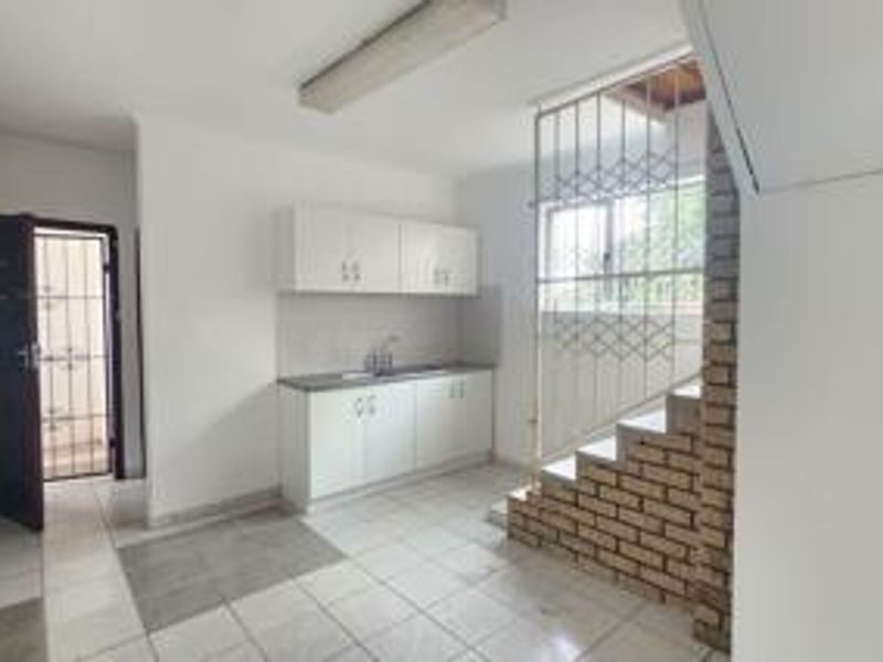 A compact, empty kitchen area featuring white built-in cabinetry, a stainless steel sink, tiled flooring, and a distinctive brick staircase feature.