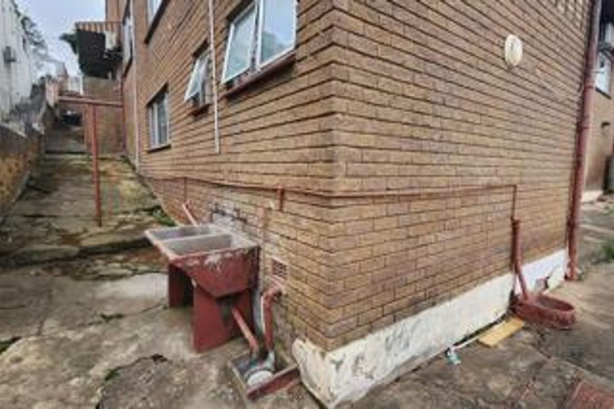 Rear exterior view of the property showing brick facade, concrete paving, and an external laundry sink area with visible signs of wear.