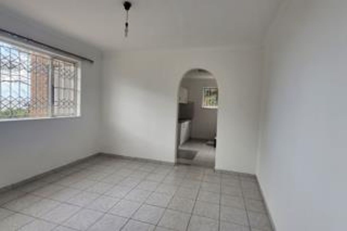 An empty living room featuring tiled flooring, a large window with security bars, and an arched entryway leading to the kitchen area.