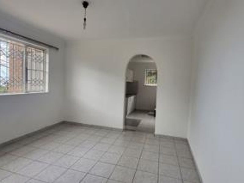 An empty living room featuring tiled flooring, a large window with security bars, and an arched entryway leading to the kitchen area.