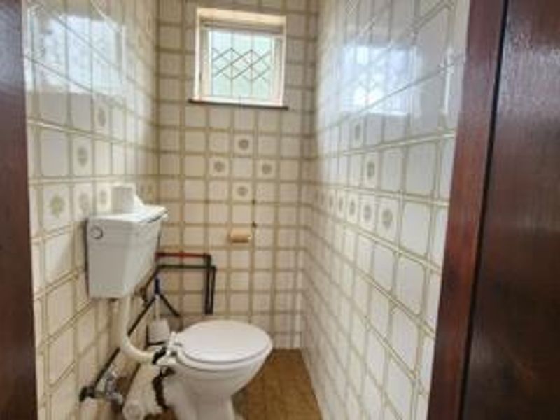 A compact, dated bathroom featuring full-height ceramic wall tiling, a standard toilet, and a small window with security bars.