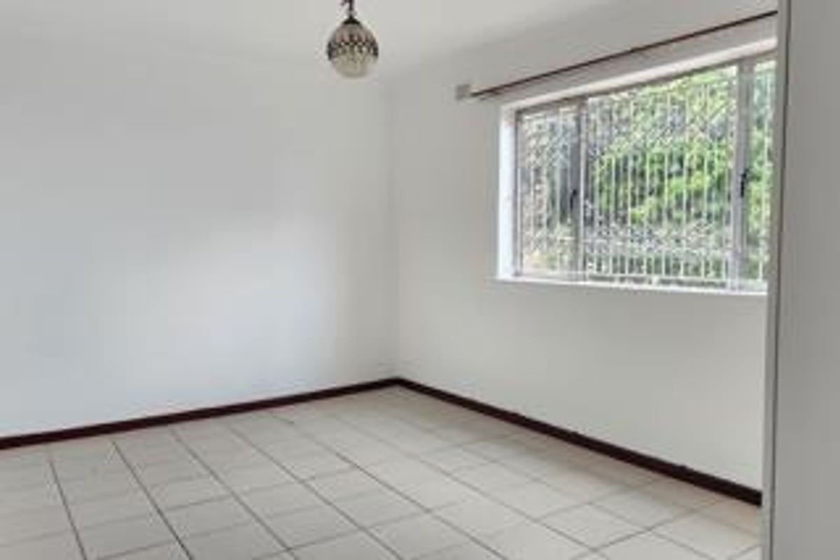 A clean, empty bedroom featuring tiled flooring, a built-in cupboard, and a window fitted with security bars.
