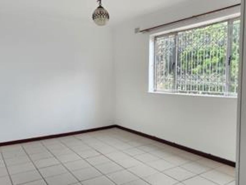 A clean, empty bedroom featuring tiled flooring, a built-in cupboard, and a window fitted with security bars.