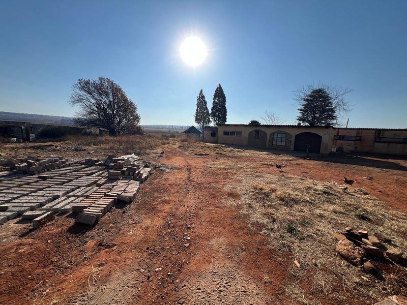 A wide-angle view of the rear exterior of a farm property, featuring a single-story building with arched architectural details and expansive unmaintained grounds.
