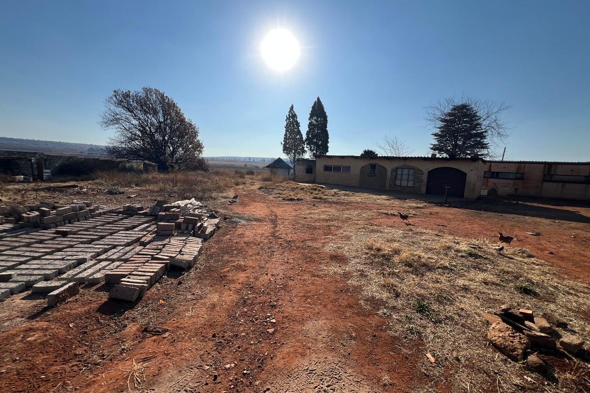A wide-angle view of the rear exterior of a farm property, featuring a single-story building with arched architectural details and expansive unmaintained grounds.