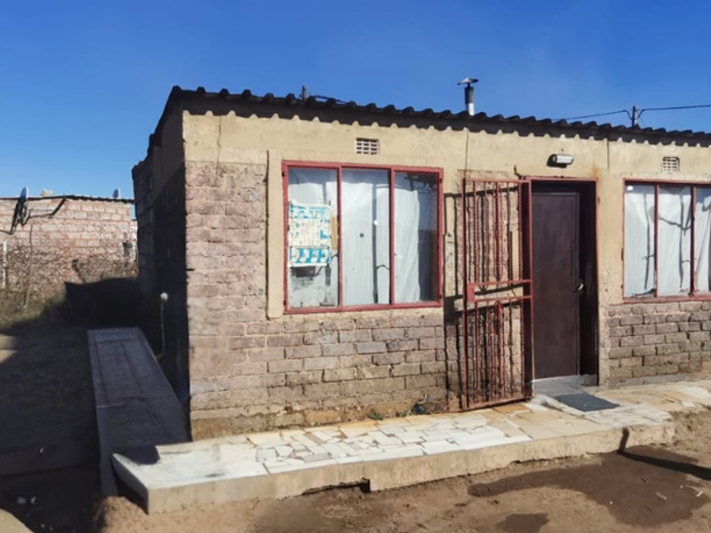 A modest single-story brick house featuring a corrugated metal roof, security gate, and tiled entrance step, situated on an unpaved plot.