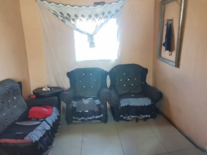 A living room area featuring tiled flooring and a window, currently showing signs of wear on the wall surfaces.