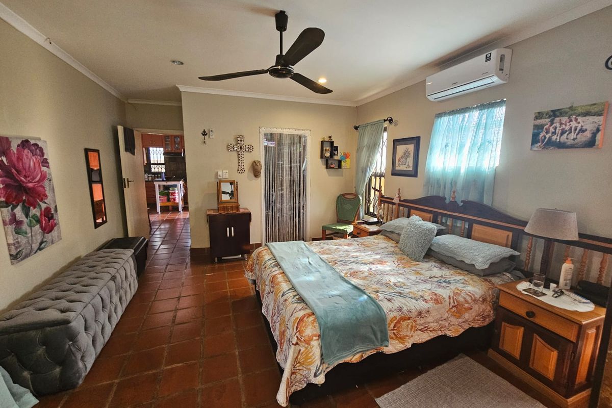 Spacious bedroom featuring terracotta floor tiles, a ceiling fan, and a wall-mounted air conditioning unit, with an adjoining view into the kitchen area.