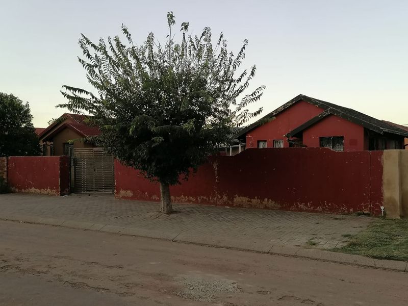 Street view of a residential property featuring a red perimeter wall, metal gate, and tiled roof structure.