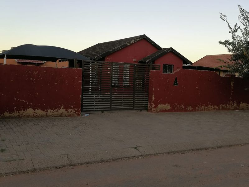 Front view of a residential property featuring a red perimeter wall, metal gate, paved driveway, and a tiled roof structure.