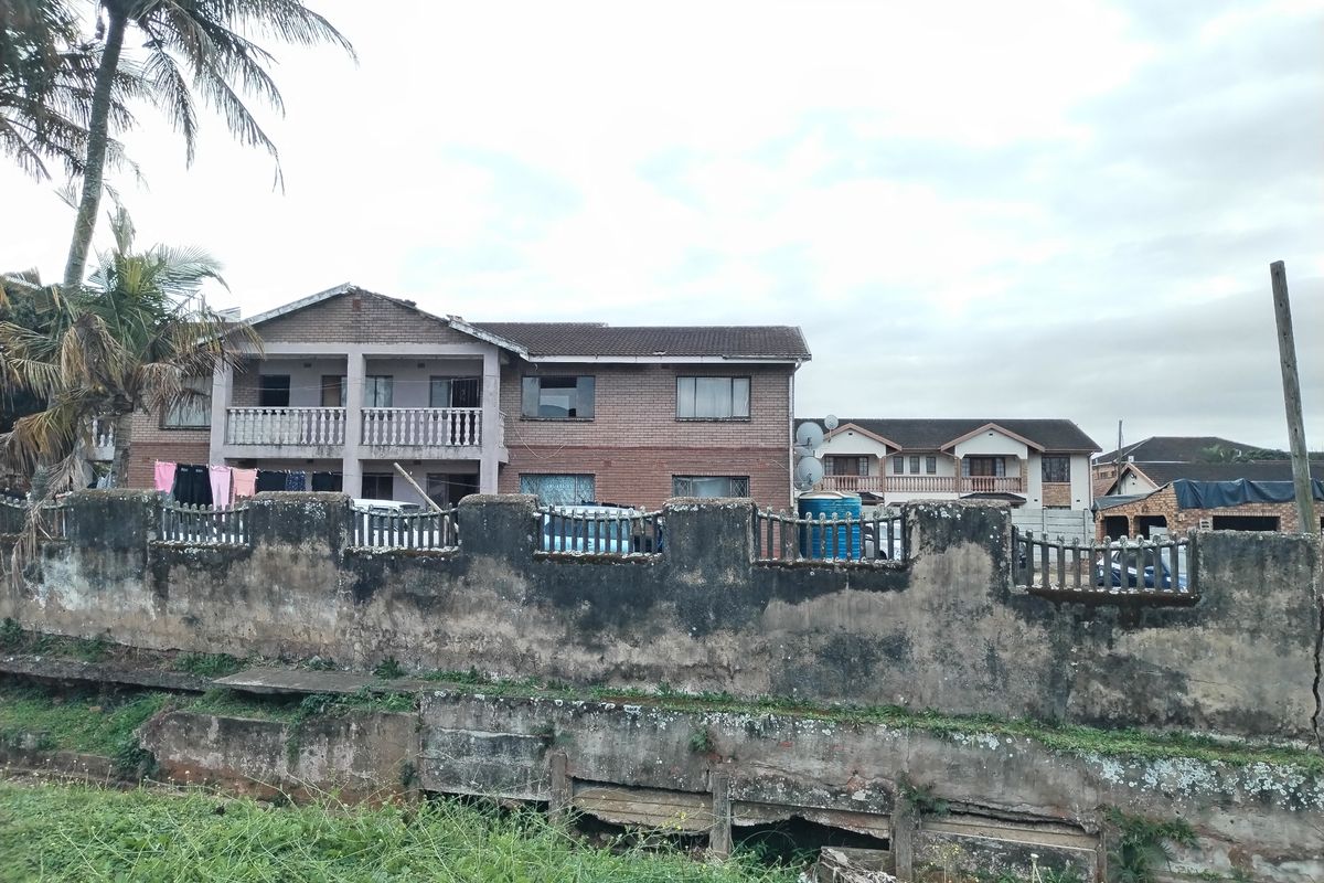 Front view of a multi-story brick residential building featuring a balcony and a weathered concrete boundary wall requiring maintenance.