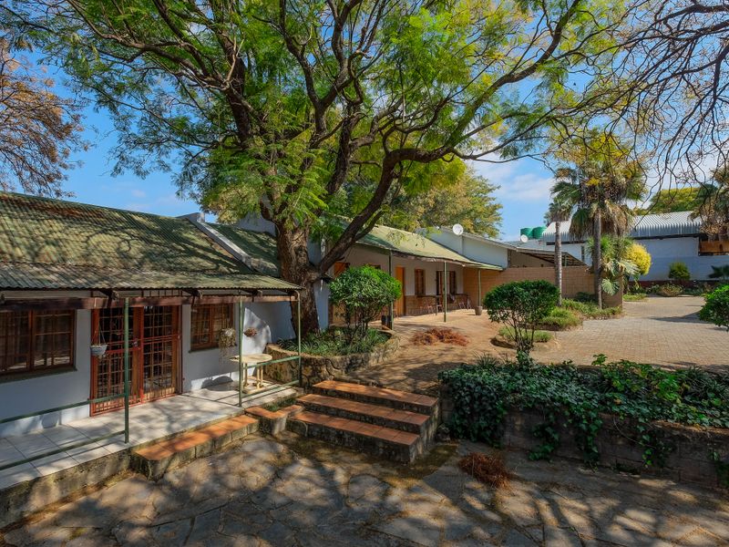 Rear exterior view featuring a paved courtyard, stone retaining walls, and covered patio areas with corrugated metal roofing.